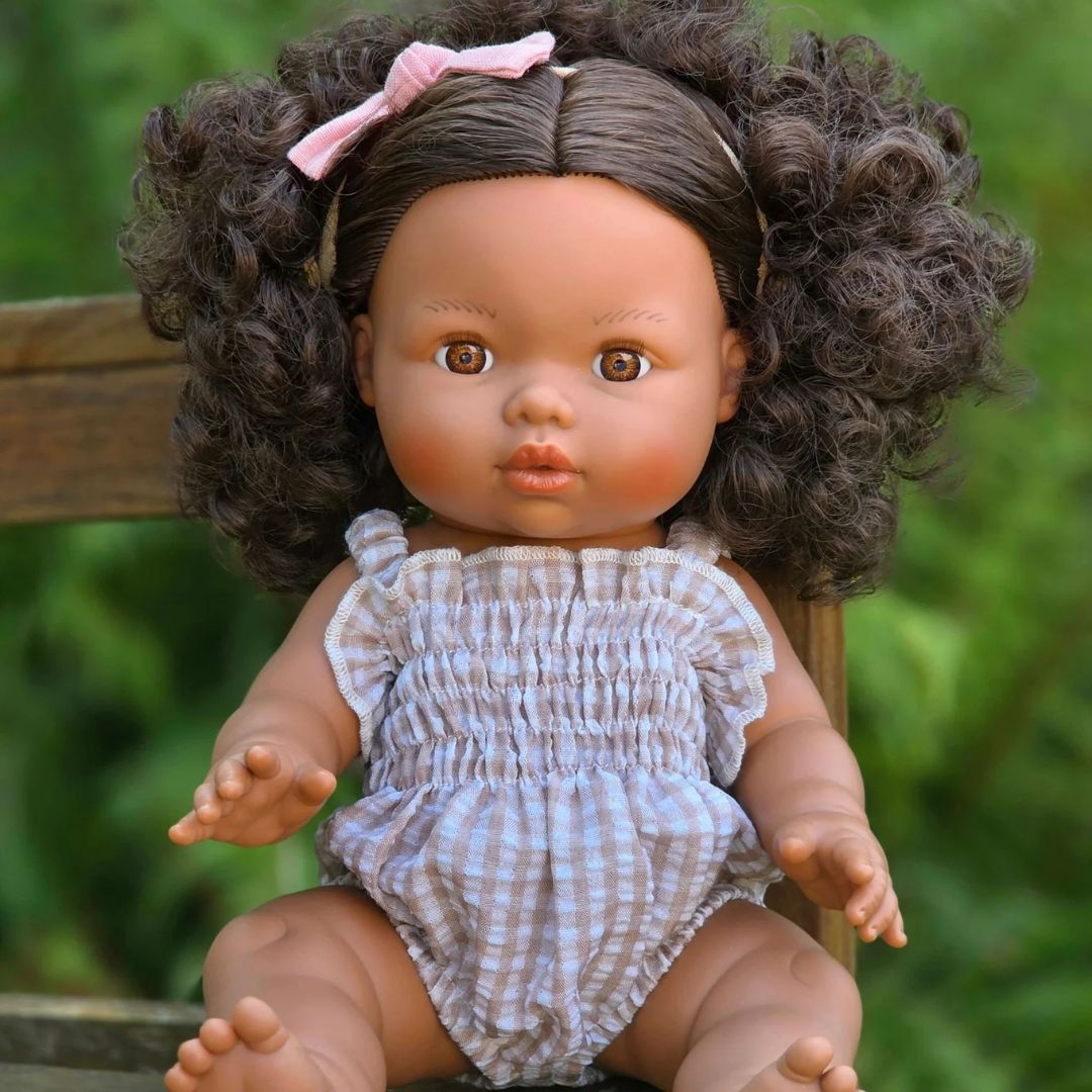 A realistic baby doll with curly brown hair, a pink bow, and a beige-and-white gingham romper, sitting on a wooden chair against a blurred green outdoor background.