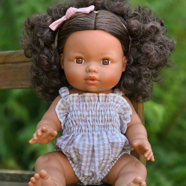 A realistic baby doll with curly brown hair, a pink bow, and a beige-and-white gingham romper, sitting on a wooden chair against a blurred green outdoor background.