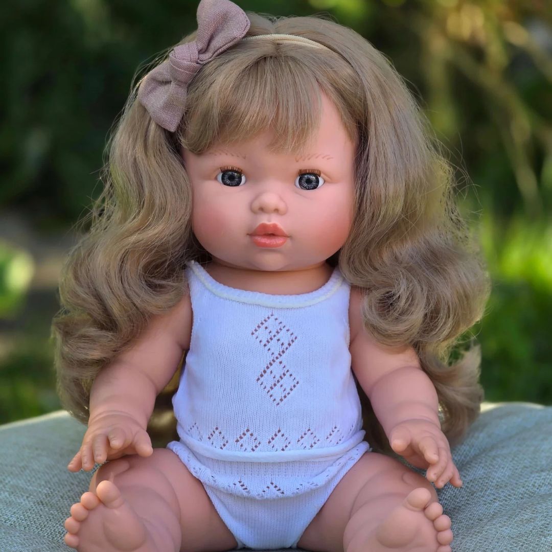 A realistic baby doll with long wavy blonde hair, a pink bow, and blue eyes, wearing a light blue sleeveless romper, sitting outdoors on a green cushion.