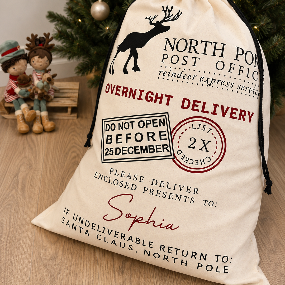 A cream-colored fabric Christmas Santa bag with black and red printed graphics, including a reindeer, 'NORTH POST OFFICE', and a personalised name 'Sophia', displayed on a wooden floor next to holiday decorations.