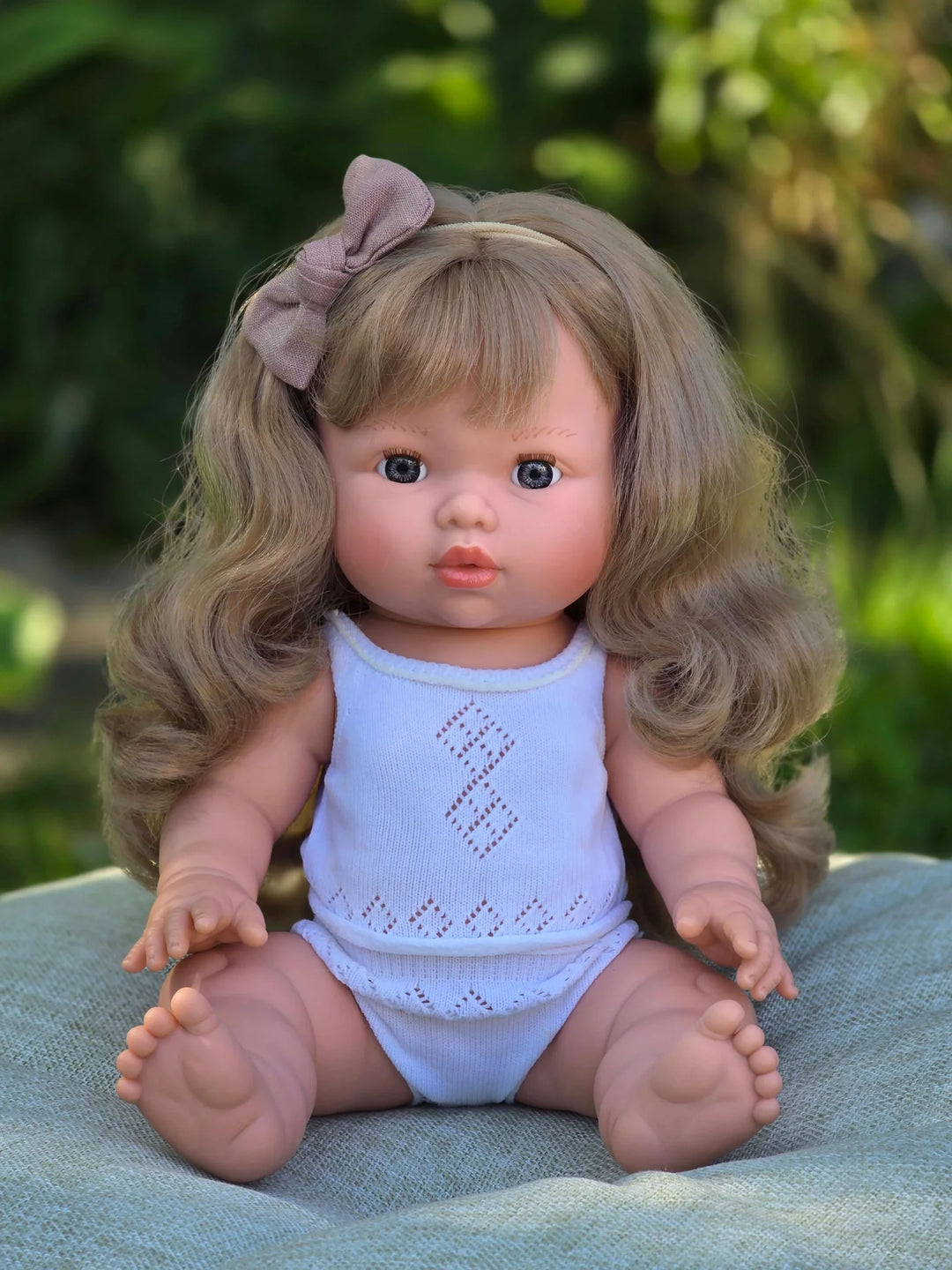 A realistic baby doll with wavy blonde hair, a dusty pink bow headband, and a white knit sleeveless bodysuit, sitting on a textured green blanket with a blurred green garden background.