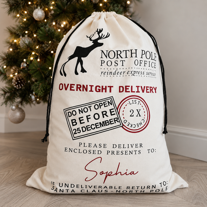 A personalised Christmas 'North Pole Post Office' Santa sack in cream canvas with black and red printed graphics, placed on a wooden floor in front of a decorated Christmas tree.