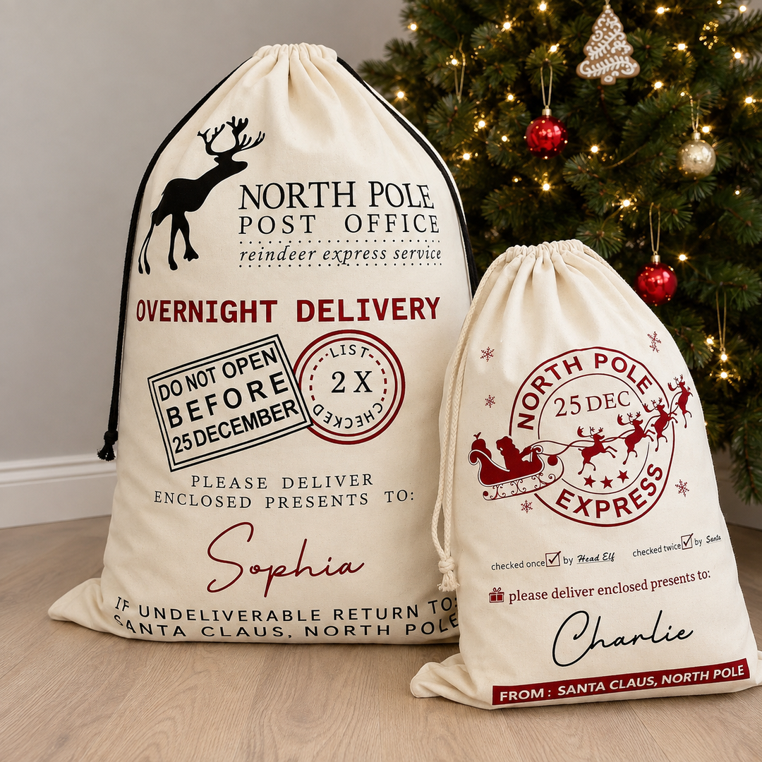 Two personalised Christmas Santa sack bags with holiday-themed printed graphics, displayed on a wooden floor in front of a decorated Christmas tree. One large cream-colored bag is labeled for 'Sophia' and one smaller matching bag is labeled for 'Charlie'.