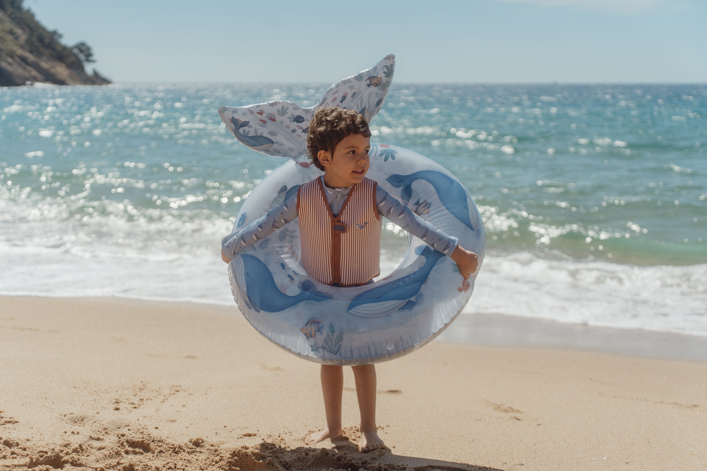 Child holding a Little Dutch whale-shaped inflatable ring on a beach with ocean in the background